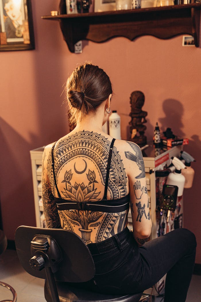 A woman with detailed body art designs sits in a tattoo studio, showcasing her intricate back tattoos.